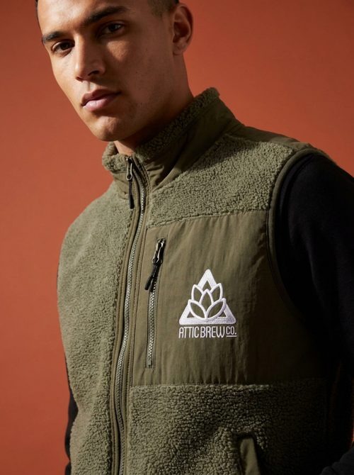 Model wearing an Attic Brew Co green sherpa vest with embroidered hops logo against an orange paper backdrop