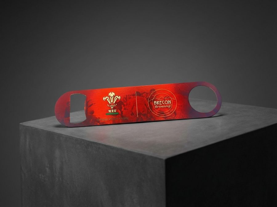 Brecon Brewing branded red metal bottle opener with WRU partnership co-brand, lit dramatically on a dark concrete plinth