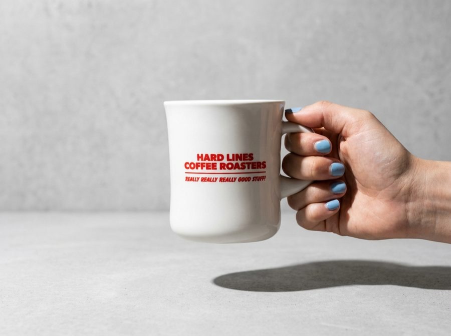 Hand with sky-blue painted nails holding a Hard Lines Coffee Roasters white ceramic mug with red printed logo against a pale concrete wall